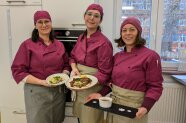 Drei Studierende zeigen Vorspeise, Hauptspeise und Nachspeise in der Schulküche.