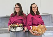 Zwei Frauen mit Kochkleidung zeigen eine Torte und Schmalzgebäck in einer Küche.