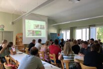 Personen sitzen an Tischreihen in einem Raum, vor ihnen Leinwand
